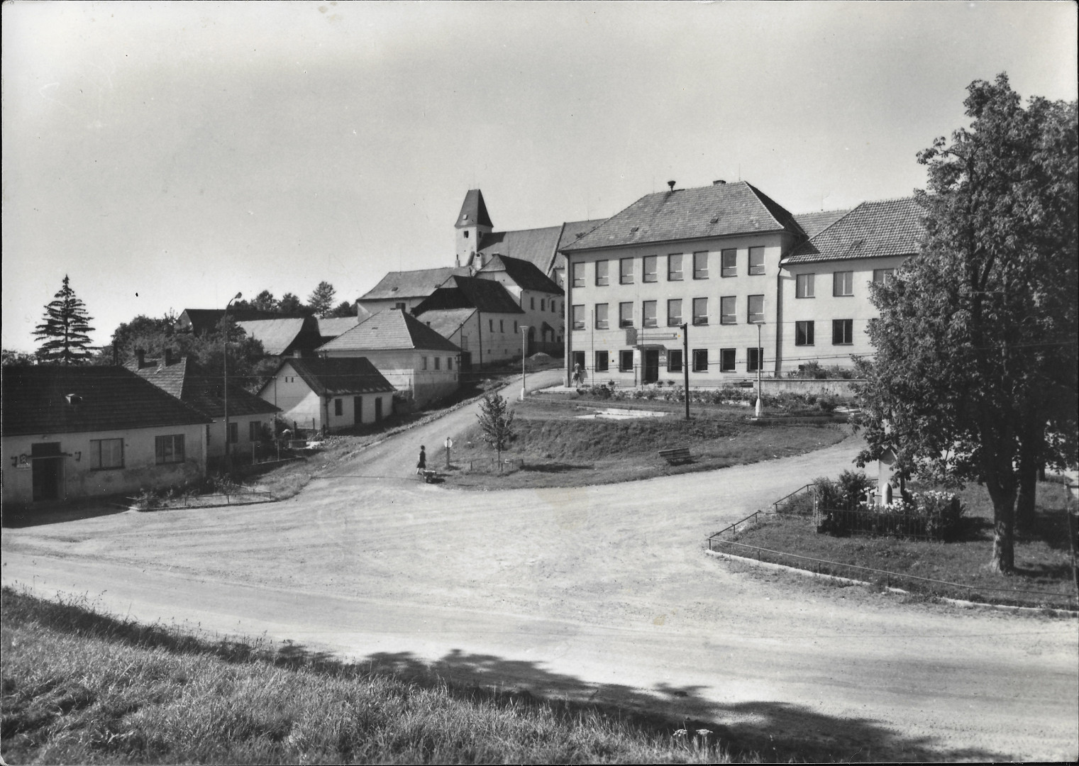 Velkou st pohlednice zabr silnice a travnat plochy nmst. V jeho horn 
      sti je kola. V prav sti je strom parku. V parku jsou ped pomnkem obt vlky
      vystaveny rapnely. V lev sti jsou domy. Z nich pln vzadu je fara a nad n kostel.