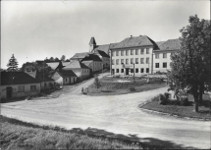 Velkou st pohlednice zabr silnice a travnat plochy nmst. V jeho horn 
            sti je kola. V prav sti je strom parku. V lev sti jsou domy. Z nich pln 
            vzadu je fara a nad n kostel.