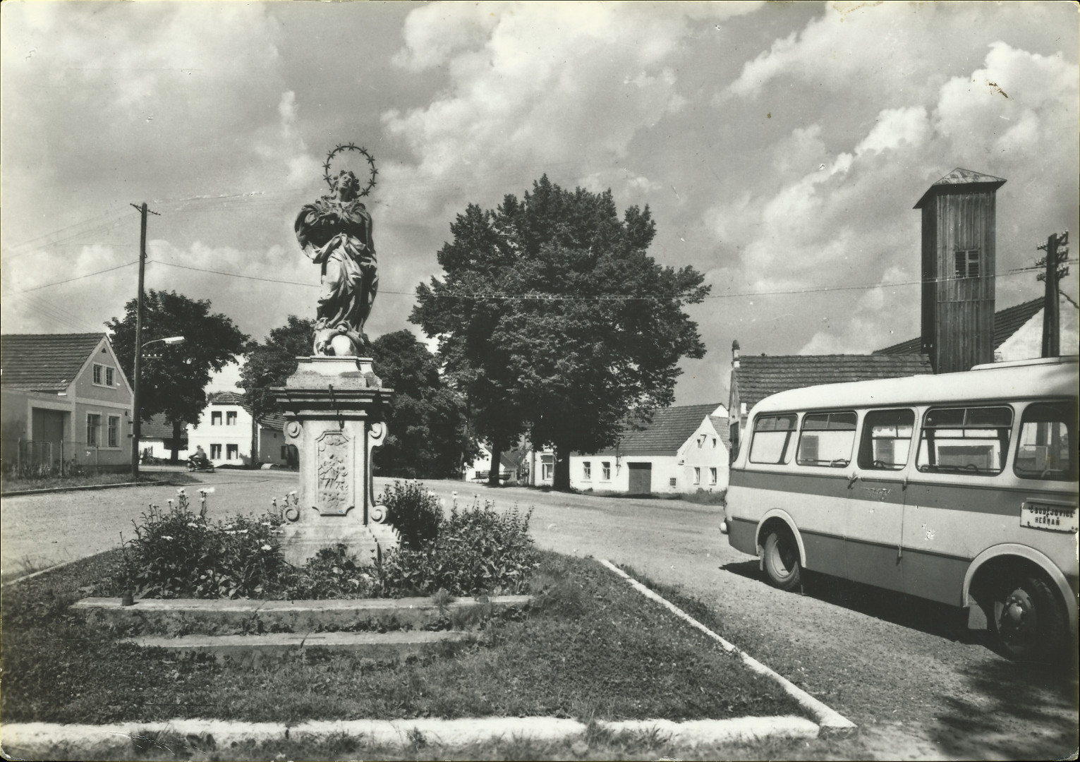 Na pohlednici je nves s
  tvercovm trvnkem lemovanm kamennm obrubnkem.
  Na trvnku je na baroknm kamennm podstavci socha Panny Marie, dobov autobus
  (tehdy se zaoblenmi hranami -aerodynamitj proveden), za autobusem je vidt
  hasisk zbrojnice (hasirna) s devnou v na suen hadic.