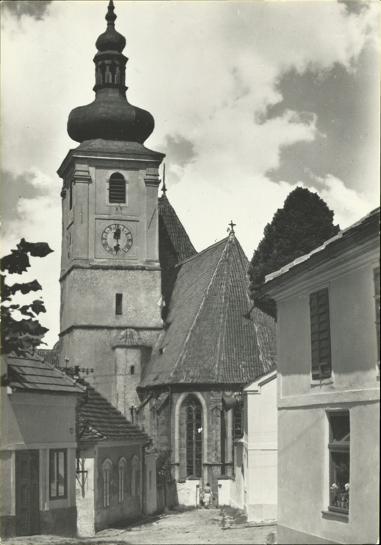 Na pohlednici je pohled na kostel piblin z jihovchodu. To znamen, e je vidt masivn v a presbyt kostela.
A za nimi vynv lo. Dle jsou vidt domy, kolem bezejmenn uliky, kterou se jde ke kostelu z Kozinova nmst.
Vedlejm prchodem na hbitov prochz ena. Na rohu jednoho domu je vidt reproduktor dobovho rozhlasu.