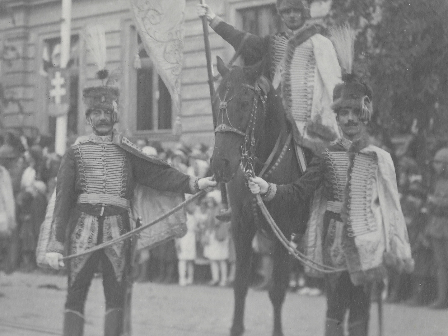 Na mstsk ulici vyzdoben znaky sten pochoduj a sten stoj ozbrojenci v uniformch po vzoru uherskho nrodnho kroje. 
          Me jt o jzdn, husarskou, ale i o p jednotku ozbrojench m욝an. Zcela urit nejde o jednotku rakousko-uhersk armdy. 
          Rakousko-uhersk armda mla toti uniformy mnohem stzlivj. Na tto polovin fotografie dr dva z nich kon, na kterm sed jezdec 
          s praporem. Maj hust rovan tmav dolmany a dlouh svtl husarsk koky, vyvn na rukvech a pedevm mohutn vyvn na 
          stehnech tmavch kalhot. Na hlavch maj tmav vlcov koeinov epice s tmavmi bambulemi a svtlmi chocholy. Nalevo od bambul maj
          jet tmav peka. Po stranch epic maj svtl aplikace (nejspe zakonuje st viditeln pytlk), kter zakrvaj lev spnky. 
          Uprosted epic jsou odznaky.