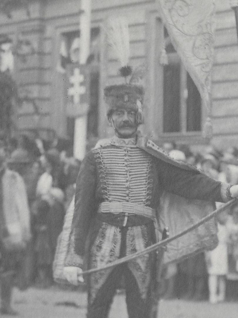 Na mstsk ulici vyzdoben znaky sten pochoduj a sten stoj ozbrojenci v uniformch po vzoru uherskho nrodnho kroje. 
          Me jt o jzdn, husarskou, ale i o p jednotku ozbrojench m욝an. Zcela urit nejde o jednotku rakousko-uhersk armdy. 
          Rakousko-uhersk armda mla toti uniformy mnohem stzlivj. Na tto sti fotografie zachycen jenom nejlpe zaosten pslunk
          jednotky. M hust rovan tmav dolman a dlouh svtl husarsk koek, vyvn na rukvech a pedevm mohutn vyvn na 
          stehnech tmavch kalhot. Na hlav m tmavou vlcovou koeinovou epici s tmavou bambul a svtlm chocholem. Nalevo od bambule m
          jet tmav peko. Po stran epice m svtlou aplikaci, kter zakrv lev spnek. Uprosted epice je odznak.