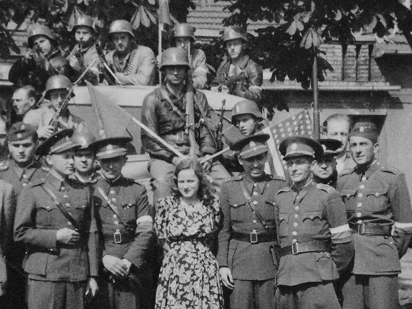 Na fotografii bez popisu stoj skupina lid uniformch prvorepublikov eskoslovensk armdy i v civilnch odvech.
  Na kraji stoj chlapec v kraasech a uprosted ena v pestrobarevnch atech. Za nimi je nejspe mal nkladn auto, na
  nm jsou mui s nmeckmi plbami a se zbranmi. Na plbch maj st rozeznateln oznaen eskoslovenskho sttu.
  Nkte maj lodiky, jin epice se ttkem a s kruhovm dnkem. Jeden m jezdeck kalhoty a holnky. Zpravidla maj puky, ale je vidt
  i kulomet s psem nboj.
  Vtina z postav na fotografii m rukvov psky s blm, ervenm a modrm pruhem. Jednomu z nich je vidt pln pepychov.
  Jeden z ozbrojenc sedcch na kapot auta dr vlajky osvoboditel tj. Sovtskho svazu a USA. Skupina je na trvnku
  a na aut. Za nimi jsou mlad listnat stromy, nejspe katany, silnice chodnk a domy.