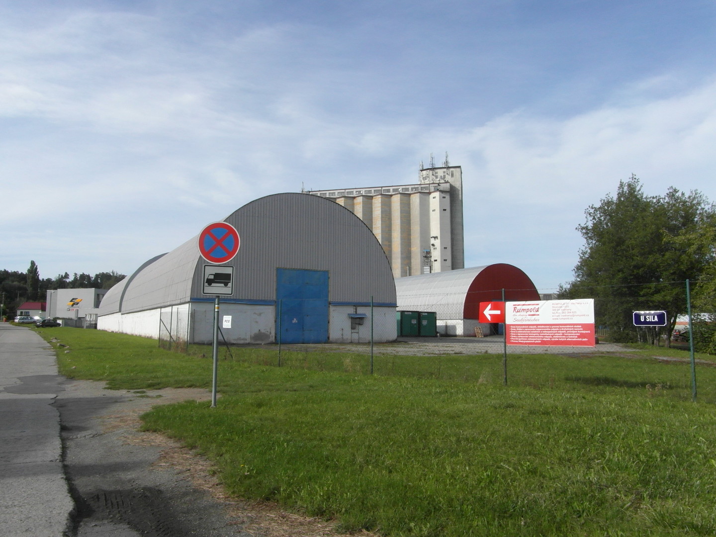 Na zbru fotografie z vletu je v pozad za dvma halami s vlcovmi stechami z vlnitch plech silo dostavn roku 1975. 
          Je vidt okraj silnice, trvnk, dopravn znaky, stromy a blo erven firemn pouta. Jedna hala m velk modr plechov vrata 
          a druh tmav erven prel.