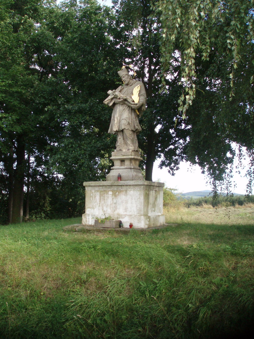 Na fotografii je na travnat vyvenin dobe osvtlen socha. Je vidt zdn 
  i kamenn podstavec. Na podstavcch jsou svky a dole je betonov truhlk s rostlinami.
  Jde o Sv. Jana Nepomuckho. K tlu tiskne k s Jeem Kristem. Je vidt i palmov
  ratolest. Svatoz s hvzdami na tomto snmku tm nen vidt. V pozad je les 
  a na obzoru hory.