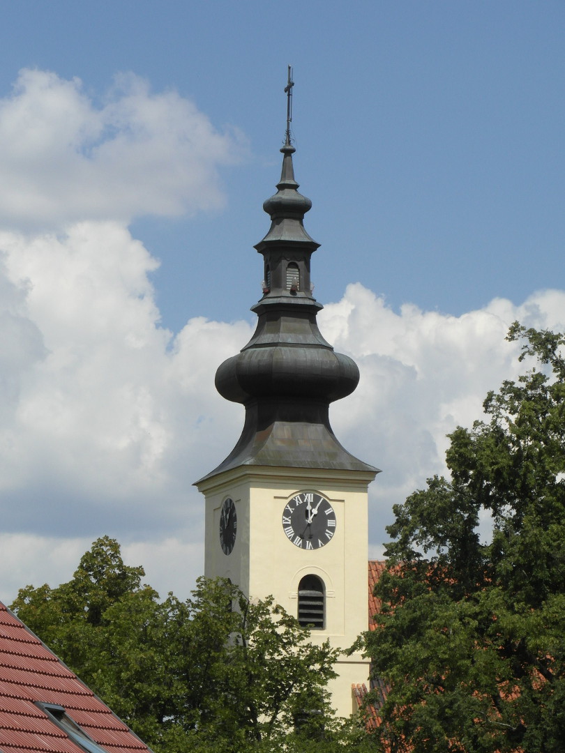 Nad stromy vynv horn st ve kostela. Na vrcholu plechov stechy ve je k s hromosvodem. Pod nm pod malou bn 
  jsou na vechny svtov strany okna se aluziemi a varovn svtla. Pod tm je velk b a roziujc se navzn plechov stechy 
  na zdnou st ve. V horn sti zdn sti ve jsou hodiny s ernm cifernkem. Ozdobn rafiky a msk slice maj stbitou
  barvu. Pod cifernkem je zprava napojena lo kostela, z n je vidt jen kousek, kter je jet k tomu zsti zakryt stromy. Pod
  hodinami je vt okno se aluziemi a o patro n dal, kter je zsti zakryto stromem, kter dole zakrv levou st ve. Vlevo
  dole je jet st stechy domu. Doln st ve je mimo zbr.