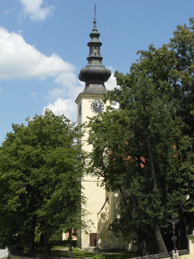 Na fotografii je vidt v kostela od jeho paty a ke pice vetn bn, hodin a sten stromem zakrytho okna. st ve 
  kryj stromy. Je vidt zatravnn plocha starho hbitova, vchod do ve a sti hbitovn zdi. V horn sti zdn sti ve jsou 
  hodiny s ernm cifernkem. Ozdobn rafiky a msk slice maj stbitou barvu. Pod cifernkem je zprava napojena lo kostela, 
  z n je vidt jen kus, kter je jet k tomu zsti zakryt stromy. Pod hodinami je tm zakryt okno a o patro n je dal,
  kter je vidt trochu vc.