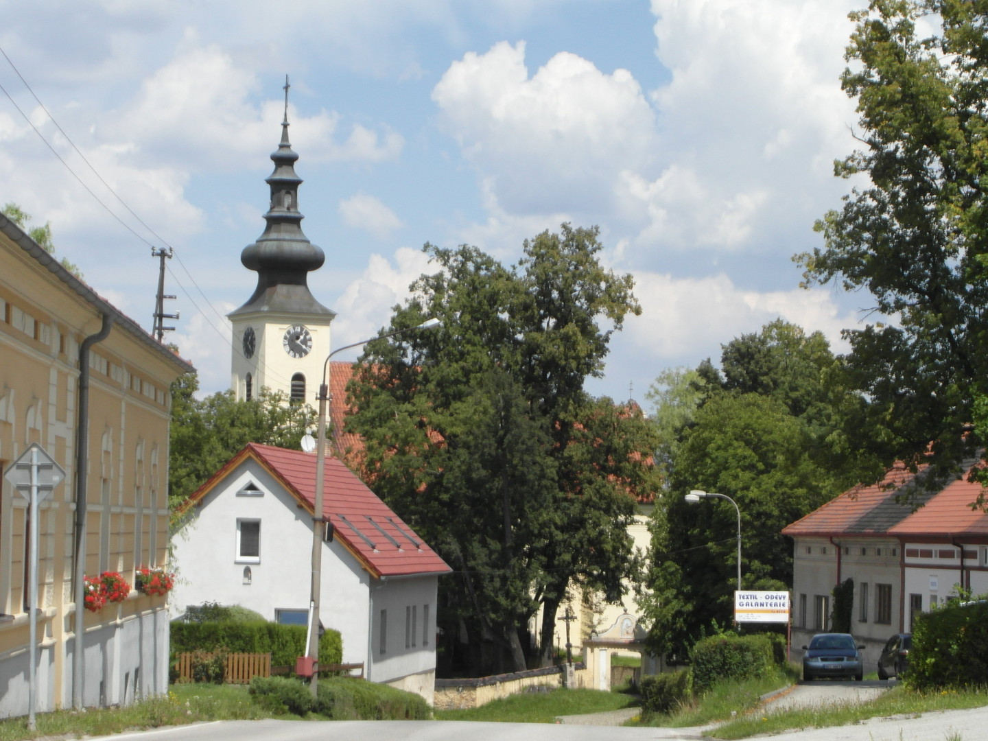 Na fotografii pozen na ulici Budjovick vedle parku (v nm jsou Bo muka) je vidt domy a velk listnat stromy,
  z nich nkter zakrvaj st lod kostela Vech Svatch. V kostela vynv nad jednm ze strom a stechou domku ped n. Kostel 
  je vyfocen zpovzdl.