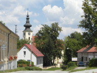 Na fotografii pozen na ulici Budjovick vedle parku (v nm jsou Bo muka) je vidt domy a velk listnat stromy,
          z nich nkter zakrvaj st lod kostela Vech Svatch. V kostela vynv nad jednm ze strom a stechou domku
          ped n. Kostel je vyfocen zpovzdl.