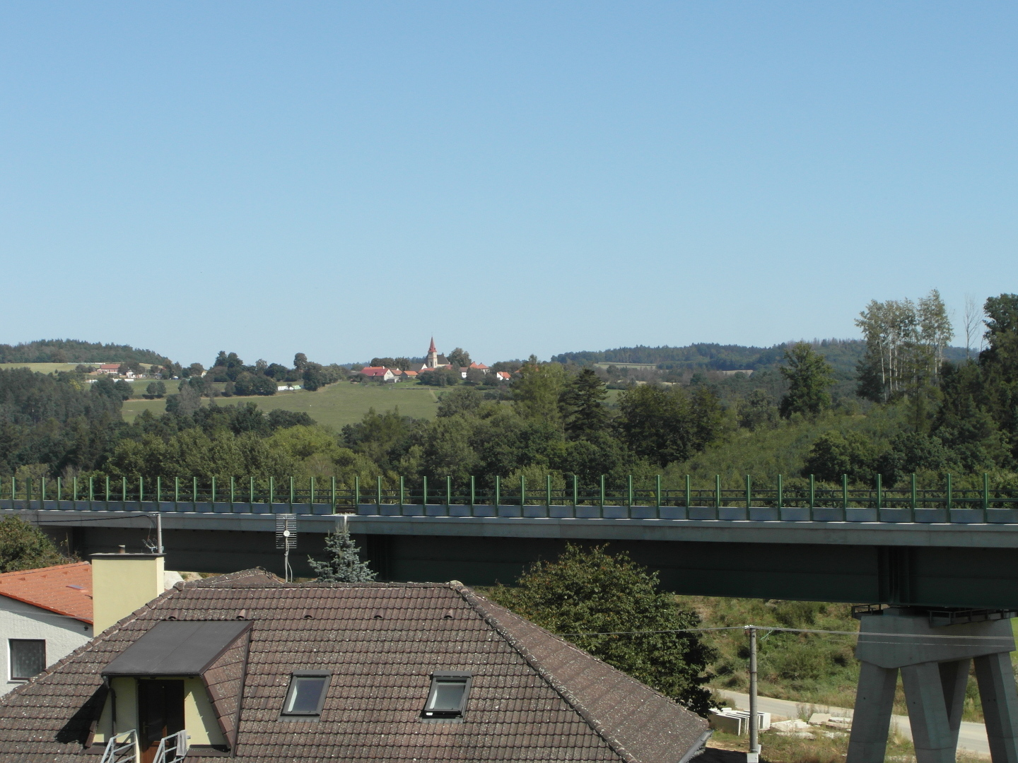 Nad stechami dom je vidt elezobetonov most s rozestavnou trat vysokorychlostn eleznice. Na obzoru je vidt krajina s kopci a vesnice
  se lutou v kostela.