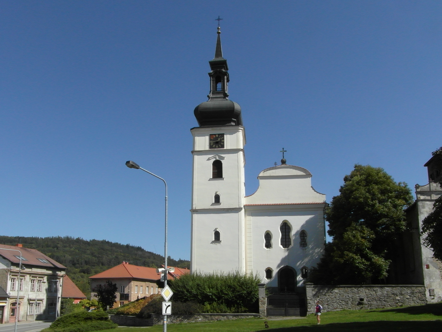 Na vyobrazen je krsn ble naten barokn kostel s barokn chrmovou lod, baroknmi okny a plechovou bn. Na fotografii je tak
  mohutn listnat strom, louka, domy a v pozad zalesnn vrch.