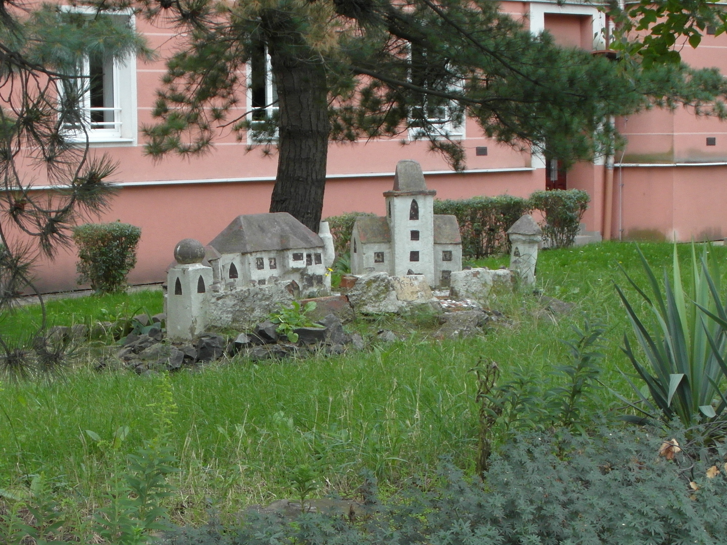 Miniatura hradu m ed stny a ern okna. Na stechch jsou vidt nznaky erven barvy. Hlavn tybok v se sedlovou stechou, piblin uprosted
  fotografie vystupuje trochu pedsunut z palce. Nalevo od n je jet jeden velk palc s komnem, kter vystupuje z rohu bon stny a je ohnut smrem
  nahoru. Palc je opaten arkem. pln vlevo je mal tybok vika s kulatou stechou. Za n je v zkrytu men palc. Napravo je vlcovit vika
  s kuelovou stechou. Ped hradem je zhon, kolem nj trva a za nm roste borovice. Dm v pozad m rovou omtku.