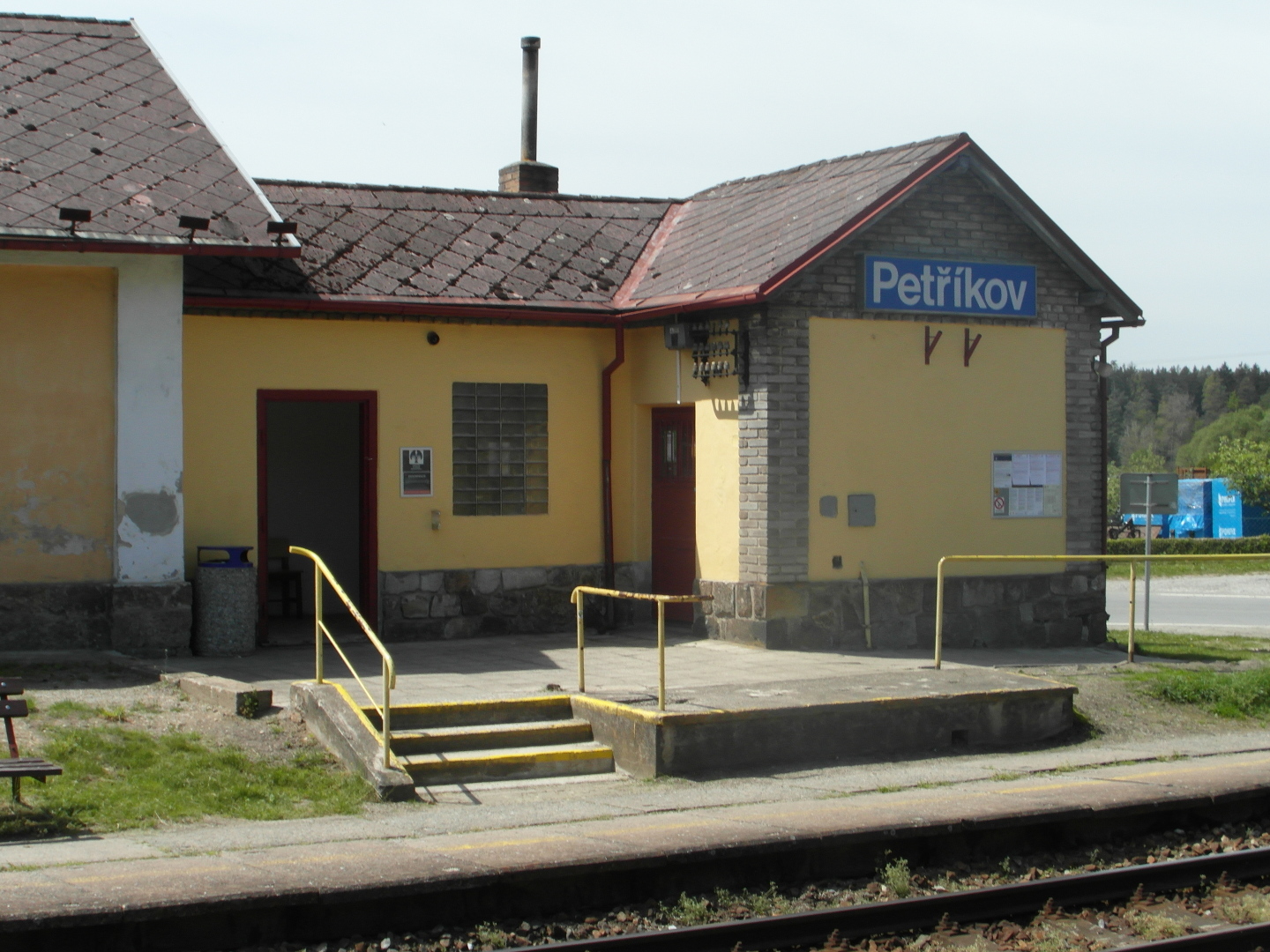 Na fotografii je st budovy vlakov zastvky s typickm lutm ntrem. Je vidt modrou ceduli s blm npisem Petkov, 
  kousek kolej, nakldac rampu a schody.