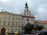 Vedle dominantn okrov budovy je na fotografii bl kostel s ervenmi oddlenmi st ploch. V doln sti je zsti zakryt
          listnatm stromem.